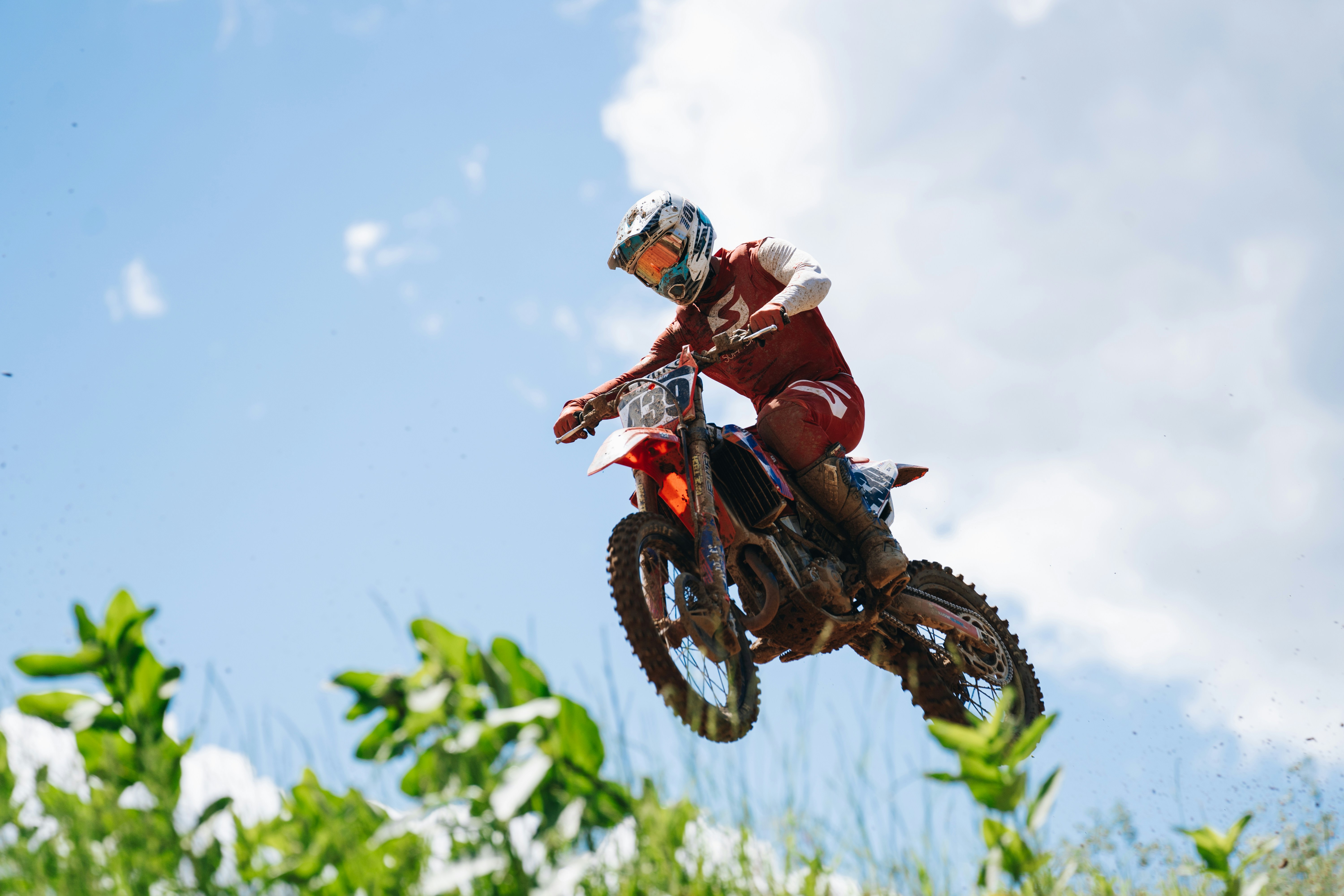 A person in red and white attire, attached to a race series motorcycle, performing an aerial stunt by jumping on a grassy ...