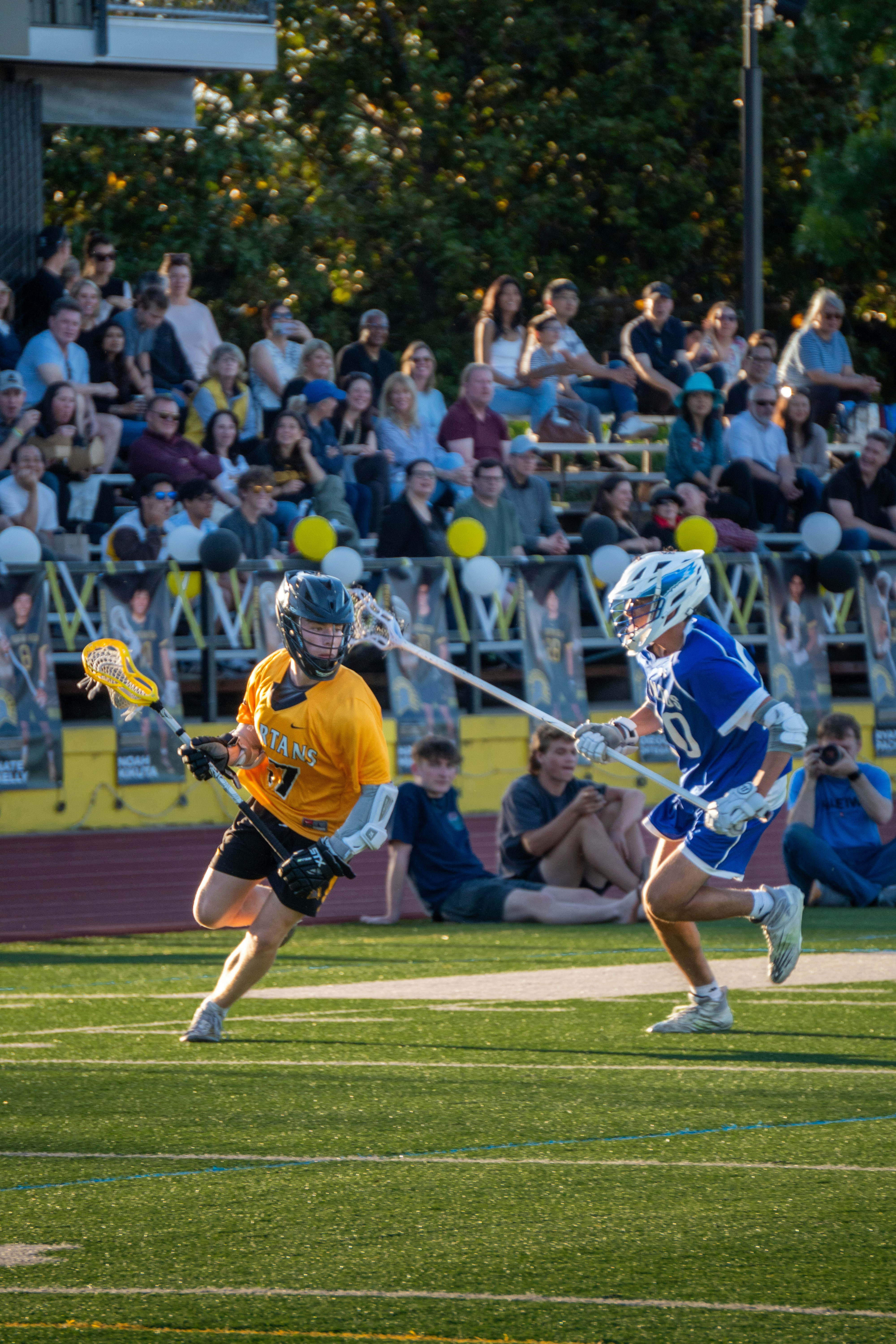 Two lacrosse players run on green turf; the player in the yellow jersey holds his stick in front and the player in the blu...