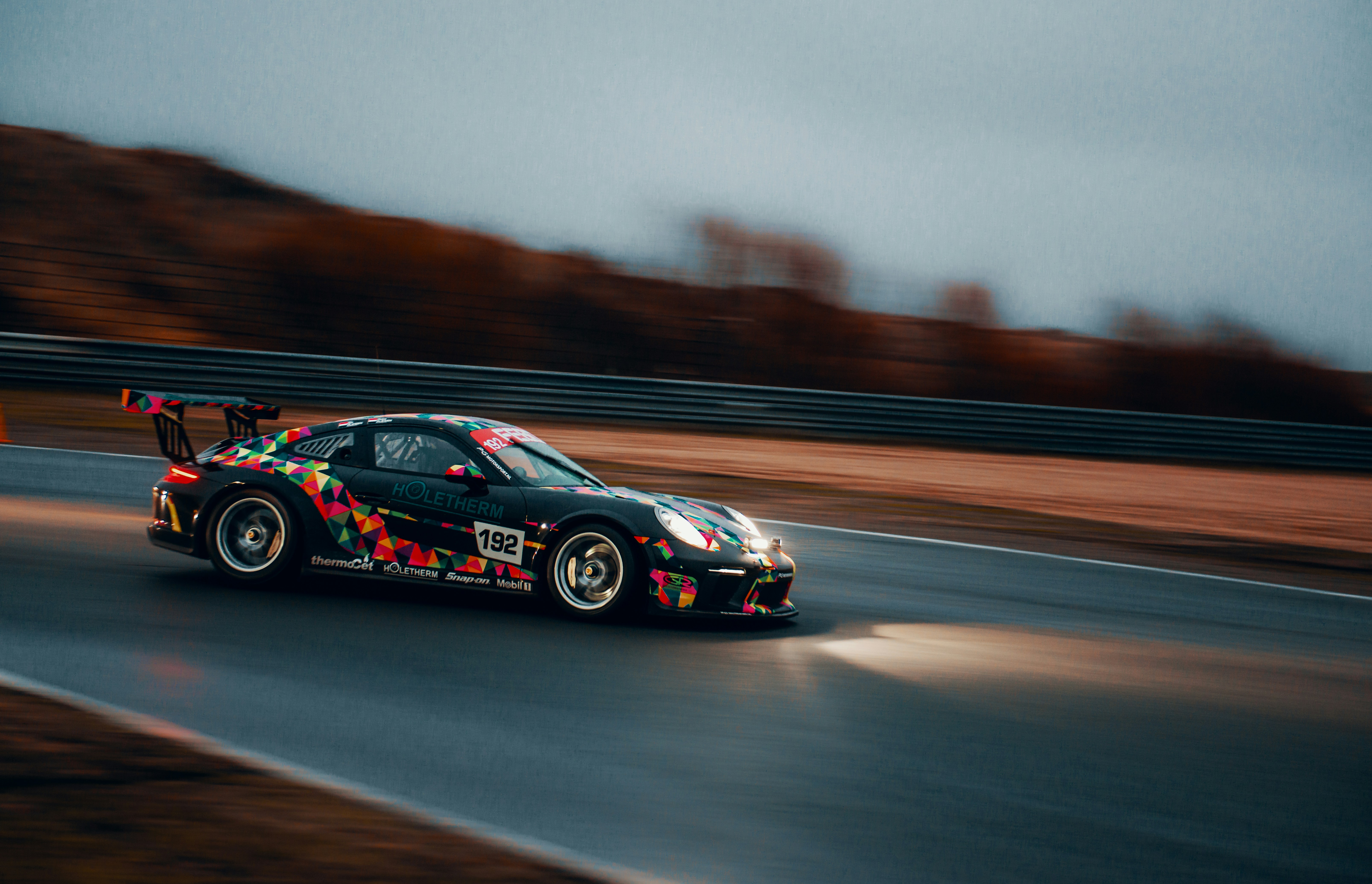 A low-to-the-ground, multicolored race car with a large spoiler and white headlights on a road track. It has a large wing ...